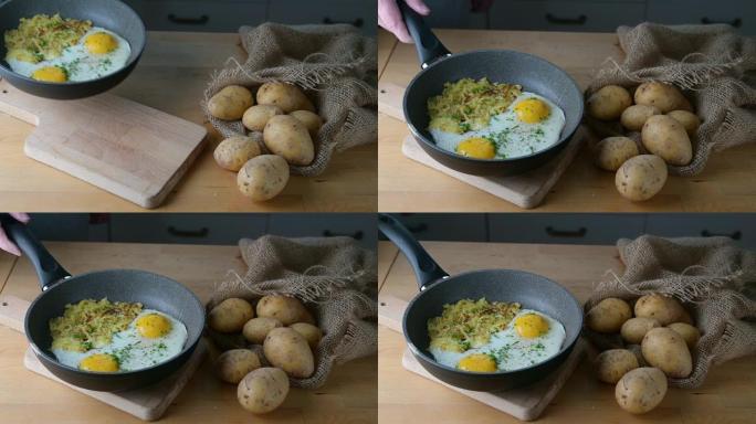 将煎蛋和瑞士烤肉 (马铃薯煎饼) 的煎锅放在木制厨房桌子上的切菜板上，上面放着一袋土豆