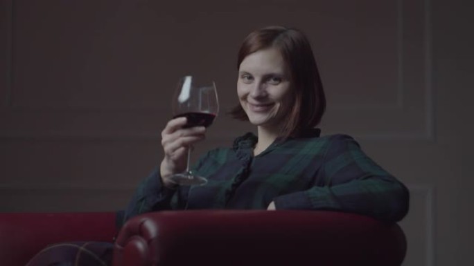30多岁的年轻女性坐在家里舒适的红色扶手椅上喝红酒。女人独自微笑着伸出手来喝杯红酒
