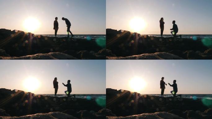 可爱的男人站在膝盖上并在日落时向女友求婚的剪影。年轻惊讶的快乐女人同意结婚，对男人求婚。日落时海滩上