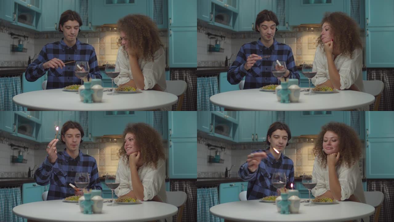 穿着休闲服装的20多岁年轻快乐夫妇在舒适的蓝色厨房里用蜡烛吃饭。男人试图以慢动作的有趣方式点燃蜡烛的