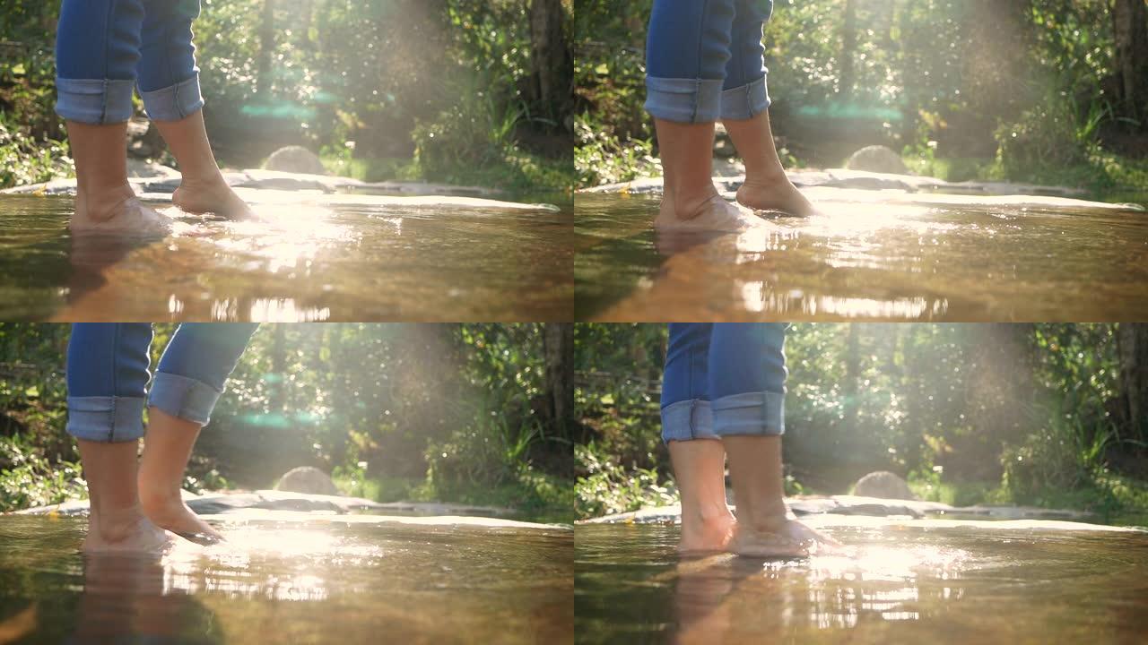 赤脚的年轻女子在缓慢流动的溪流中的水中飞溅和踢腿。女士们在瀑布享受大自然的假期。暑假生活方式的概念。