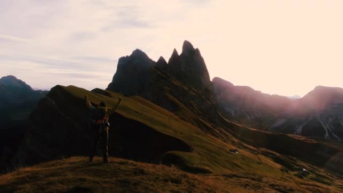 年轻人在Seceda山上徒步旅行，在日出时向山顶举起手臂。背包，黄色夹克，靴子，豆豆。前往意大利特伦