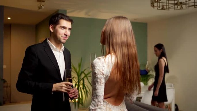 美丽的年轻夫妇在庆祝聚会上站在客厅的起泡酒上聊天和调情，他们的朋友在后台聊天