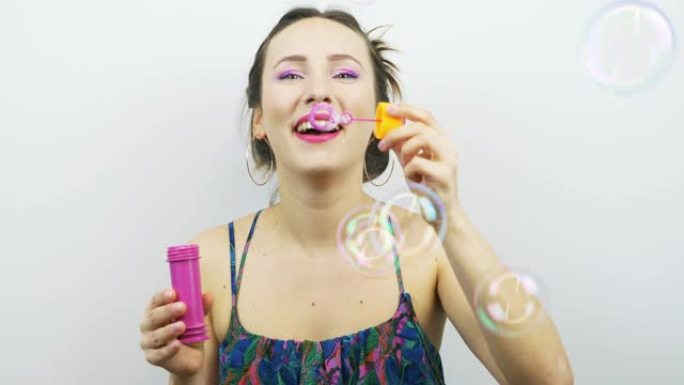 快乐的笑女孩与粉红色的化妆吹肥皂泡。滑稽微笑的女人在白色背景下吹肥皂泡。女性玩得开心