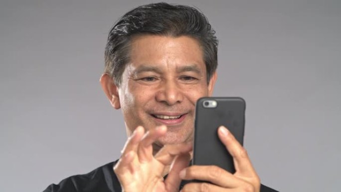 特写高级男子使用手机浏览，非常开心的微笑，孤立在灰色背景中。穿着黑色衬衫的亚洲男人。