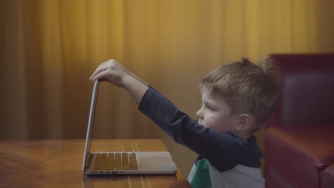 金发碧眼的学龄前男孩在笔记本电脑上打开和打字，微笑着看着电脑屏幕。孩子坐在桌旁在家在线学习。侧视图