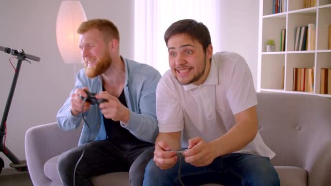 两个成年英俊的白人男子坐在室内沙发上玩电子游戏的特写肖像。获胜者取笑失败者