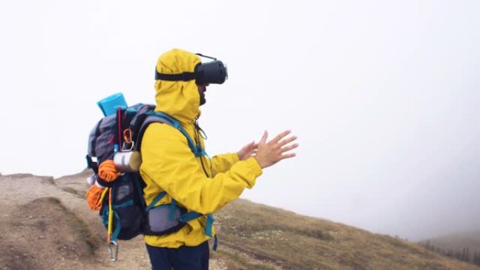 VR虚拟现实耳机。年轻的徒步旅行者在多云的天空和雾的背景下移动双手。山峰上的黄色兜帽夹克和背包。
