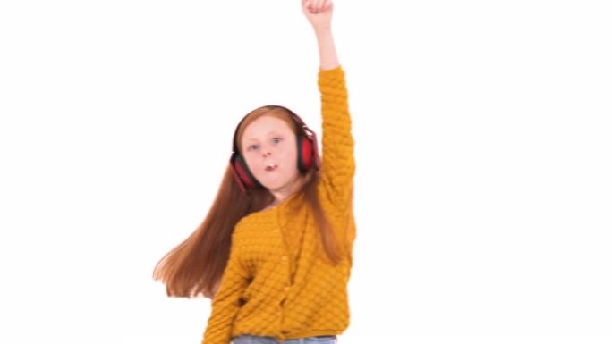 戴着无线耳机的红头发女孩听着音乐和舞蹈。可爱的年轻模特吃棒棒糖。孤立，在白色背景上