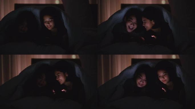 情侣亚洲少女晚上看手机，在卧室的毯子下一起聊天