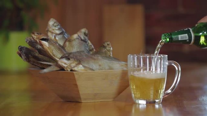 在鱼干前将一瓶啤酒倒入玻璃杯中。一杯冷新鲜啤酒放在木条上。小吃啤酒。带干鱼的木碗。
