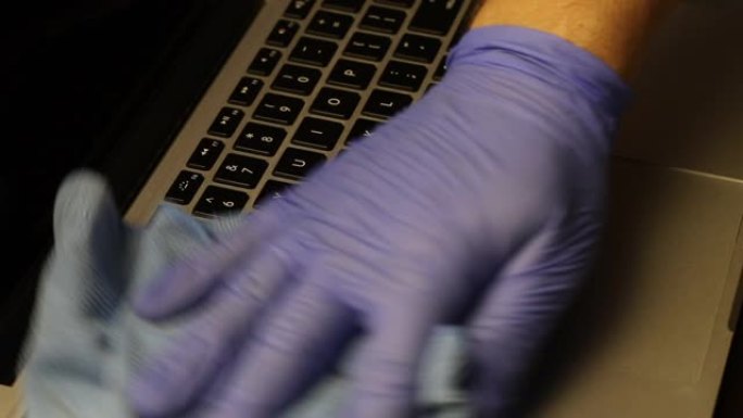 一个戴着橡胶手套的男人正在清洁他的笔记本电脑。他用餐巾纸擦拭笔记本电脑键盘。保护工作设备免受病毒感染
