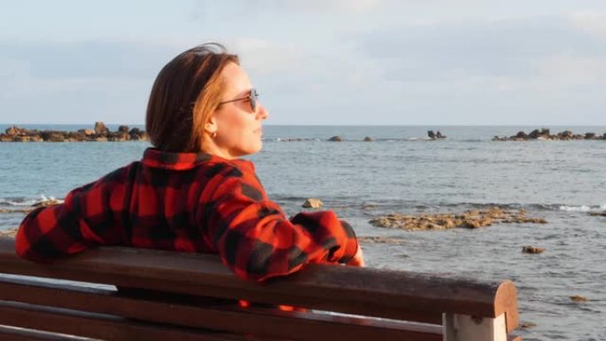 孤独忧郁的黑发女人坐在空长廊的木凳上，看着大海和日落，特写后视