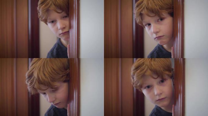 可爱的红发白人男孩的特写镜头从门口后面望出去，然后躲在后面。害怕的小孩在室内的肖像。恐惧，压力，童年