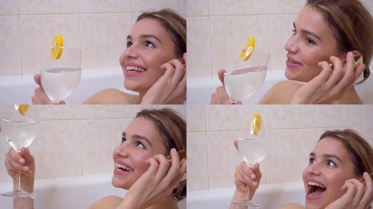 快乐迷人的女性在浴缸里打电话，喝健康的柠檬水，大笑和微笑时聊天的手机