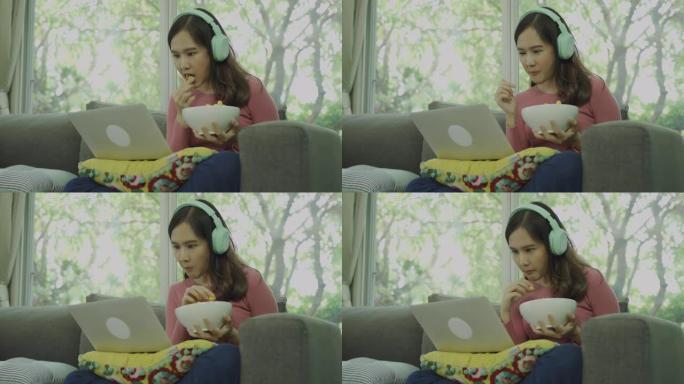 近距离拍摄亚洲年轻女子戴着无线耳机，在笔记本电脑上观看在线电影，在家吃爆米花的4k镜头。