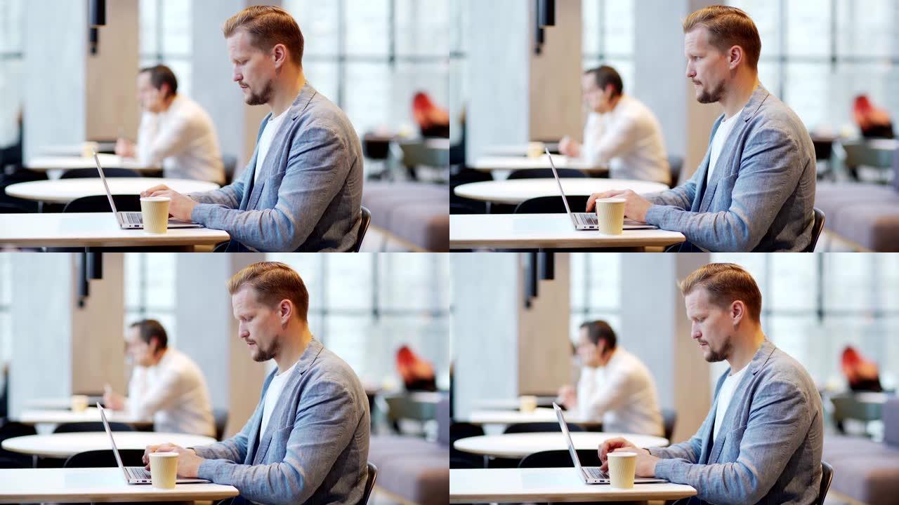 侧视中忙碌的大胡子中年商人在笔记本电脑上打字的镜头坐在咖啡馆的桌子上，散焦的商人在后台用手机聊天