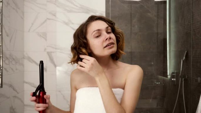 一个年轻女子的肖像，在一个大的白色浴巾造型她的头发卷发在镜子前。一个女孩洗完澡后在浴室里做发型。前视