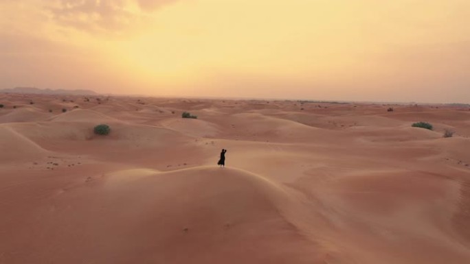 在沙漠中的沙丘上行走的一名穿着阿巴亚 (阿拉伯联合酋长国传统服装) 的妇女旁边飞行的无人机的鸟瞰图