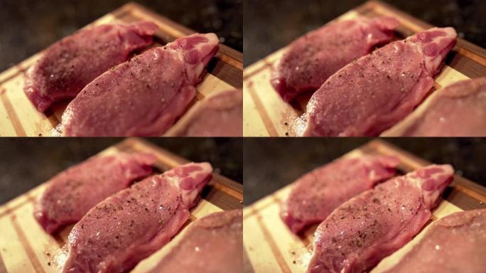 慢动作将粗胡椒粉撒在未煮熟的生去骨里脊肉猪排上，新鲜放在准备烧烤的木制砧板上