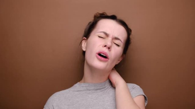 疲惫心烦的年轻女子感到僵硬疼痛颈部疼痛概念摩擦按摩紧张的肌肉患有纤维肌痛，表情阴郁，孤立在棕色背景上