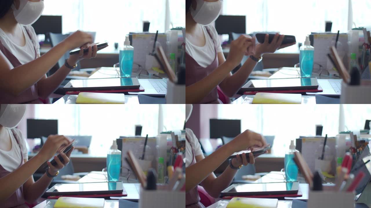 戴口罩的亚洲女商人在家庭办公室后台开始工作之前，使用酒精为基础的洗手液喷雾清洁平板电脑和智能手机。洗