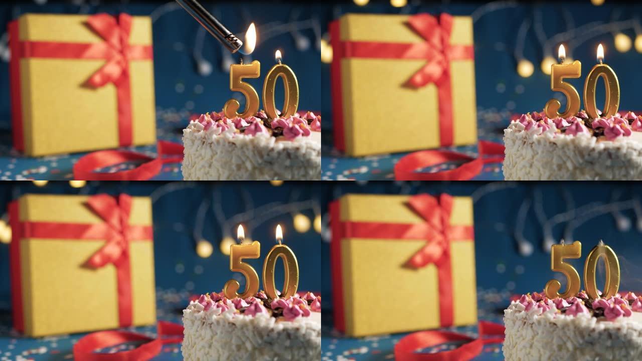 白色生日蛋糕50号点火器燃烧的金色蜡烛，蓝色背景带灯和用红丝带捆绑的礼物黄色盒子。特写