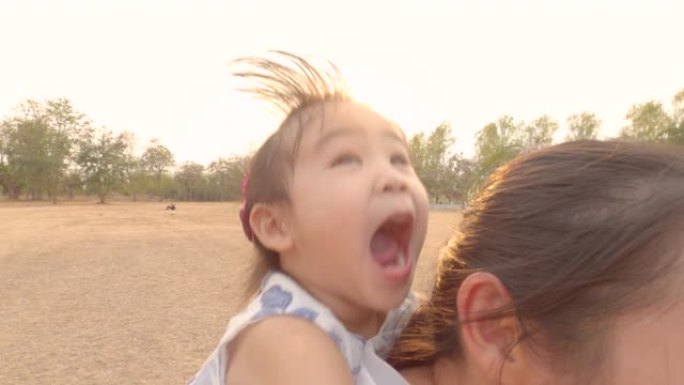 快乐的小女孩坐在公园里奔跑的母亲的背上。幸福家庭和童年的概念。