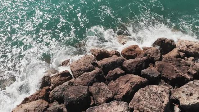 穿着红色连衣裙的女人站在岩石悬崖上，双手分开，蓝色的水撞向岩石码头海滩。海浪与岩石相撞的鸟瞰图，穿着