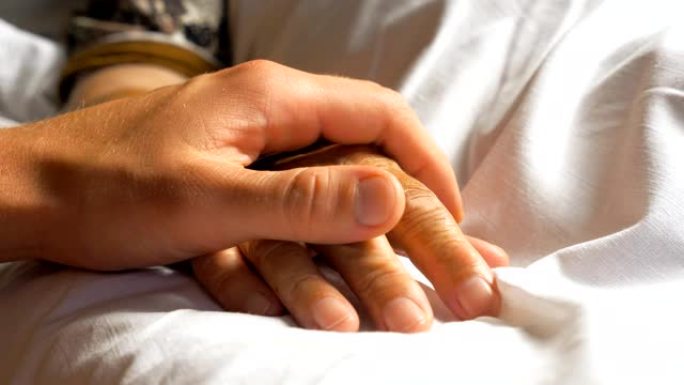 面目全非的忧心忡忡的人轻轻地抚摸着生病的祖母给予支持的手。儿子安慰躺在医院床上的年迈妈妈皱巴巴的手臂