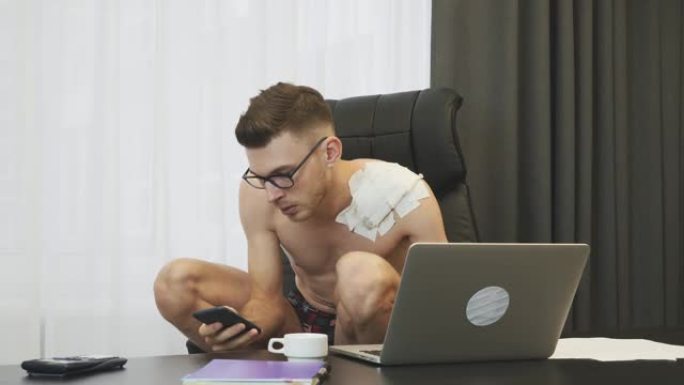 一个男人蹲在椅子上在办公室用笔记本电脑工作。穿着内衣在笔记本电脑上打字的男人。年轻的经纪人坐在自己的