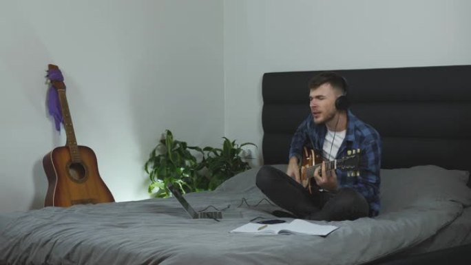 戴着耳机的男人用电吉他唱歌和弹奏。男音乐家在现代客厅的床上唱他的新歌。吉他演奏者练习吉他课。千禧一代