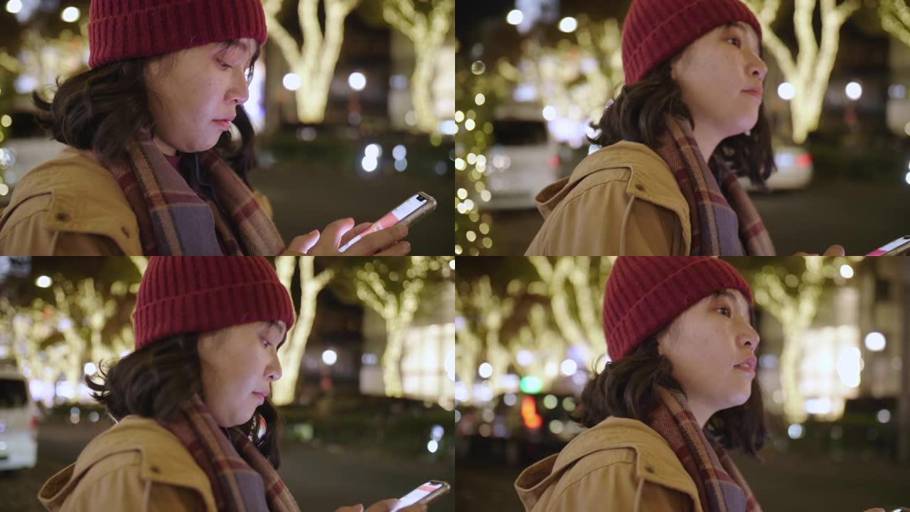 亚洲女孩女人戴上羊毛帽子和围巾，晚上在城市的智能手机上请求并等待Uber。
