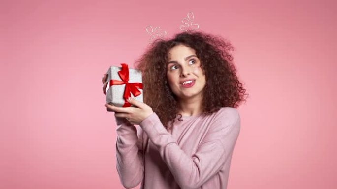 年轻可爱的女孩微笑着拿着粉色工作室背景上的礼物盒。女人在圣诞头角鹿花环。新年心情