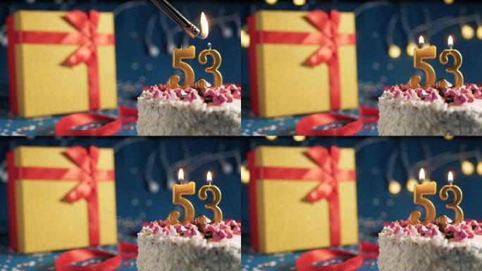 白色生日蛋糕编号53点灯燃烧的金色蜡烛，带灯的蓝色背景和用红丝带绑起来的礼物黄色盒子。特写