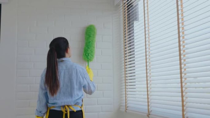 年轻的亚洲妇女打扫房子用羽毛扫帚和掸子擦拭灰尘，同时在窗户上打扫房子保持概念