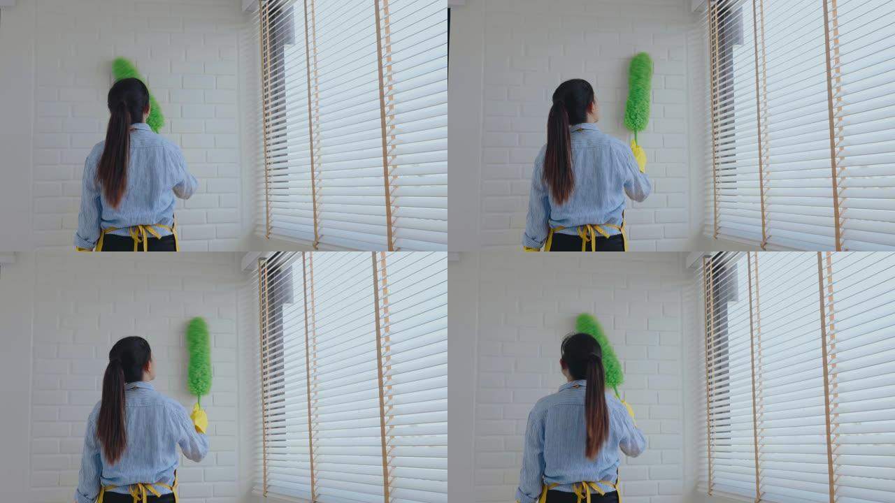年轻的亚洲妇女打扫房子用羽毛扫帚和掸子擦拭灰尘，同时在窗户上打扫房子保持概念
