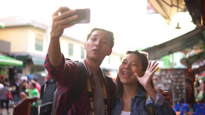 年轻快乐微笑的亚洲旅游夫妇在日落时在泰国曼谷考山路市场慢动作行走时自拍智能手机
