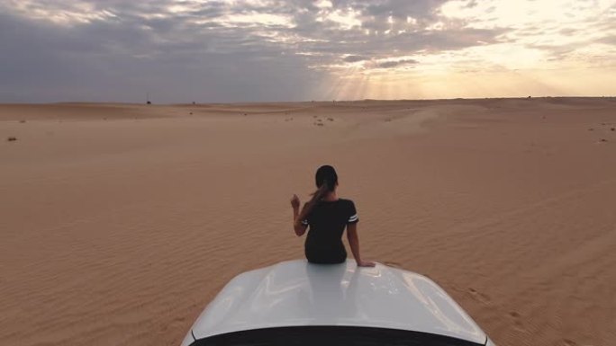 年轻的女人坐在车顶上，看着史诗般的日落，在沙漠的背景下。