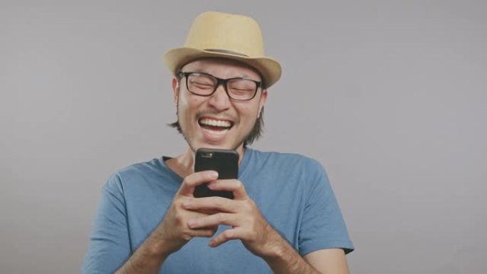 年轻的潮人看着手机屏幕，在灰色背景中孤立地笑。在镜头前戴着帽子和眼镜的亚洲男人。中枪。