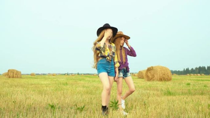 两个女牛仔走在草堆景观的乡村田野上。戴着帽子和方格衬衫的年轻奶牛女孩摆前摄像机。乡村女孩在村子里的收