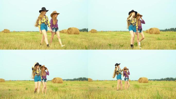 两个女牛仔走在草堆景观的乡村田野上。戴着帽子和方格衬衫的年轻奶牛女孩摆前摄像机。乡村女孩在村子里的收
