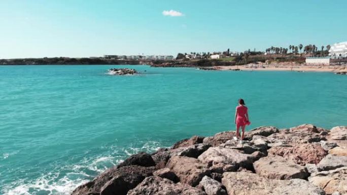 穿着红色衣服的女人在岩石海滩上行走，双手分开，看着蓝色的海洋和天空。风在拍打她的裙子。珊瑚湾塞浦路斯