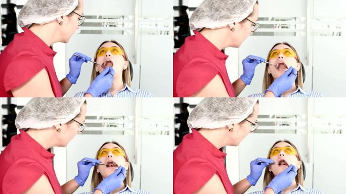 穿着黄色防护眼镜的漂亮金发女孩在踩踏医生检查她张开的嘴。女牙医在牙医仪器的帮助下检查年轻患者的口腔