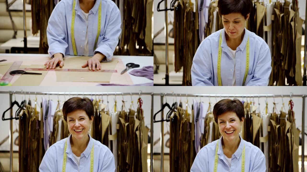 向上倾斜美丽的中年女裁缝的肖像看着相机，在制衣工作室用尺子站在桌子上做纸质缝纫图案时微笑