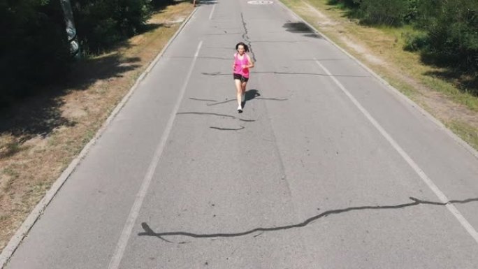 穿着粉色衬衫的漂亮苗条女孩正在练习跑步。戴着耳机的年轻黑发女人在炎热的夏日在公园锻炼