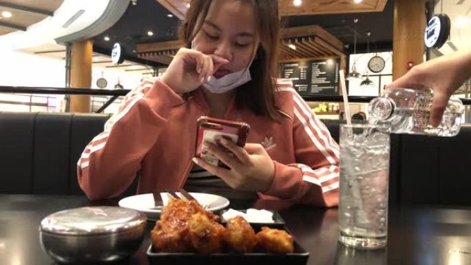 戴着口罩保护的年轻亚洲女性从饮食中休息，在餐厅吃饭，从智能手机上寻找媒体
