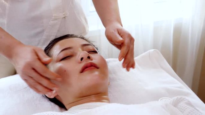 亚洲女性在美容院中心健康水疗按摩治疗，女性按摩师疗法轻轻按摩太阳穴的头部。躺在床上的女孩感到轻松舒适