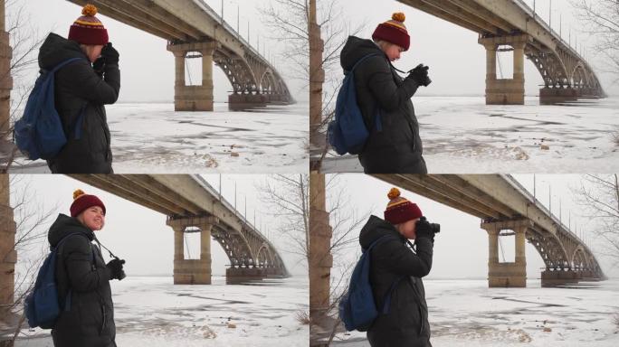 女孩游客拍摄桥下河边的冬季风景。女摄影师在大自然中拍照。