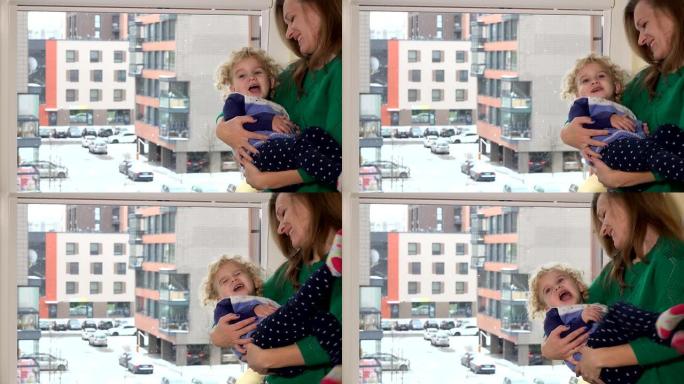 慈爱的母亲在窗户附近摆着淘气的女儿女孩。降雪